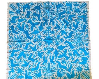 May include: A white fabric tablecloth with a blue embroidered design featuring stylized birds, flowers, and plants. The design is symmetrical and creates a repeating pattern.