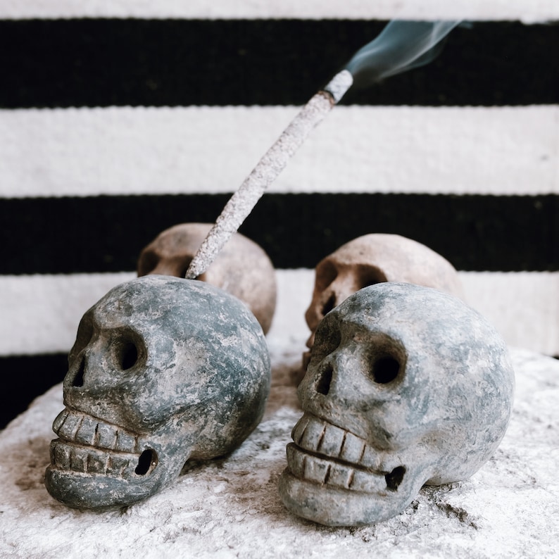 May include: Four grey and brown skull shaped incense holders with a stick of incense burning and releasing smoke. The incense holders are sitting on a white surface with a black and white striped background.