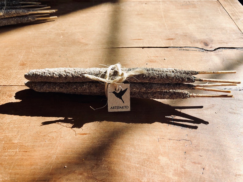 May include: A bundle of grey incense sticks tied together with twine and a brown paper tag that says "ARTEFAKTO" with a black hummingbird graphic.