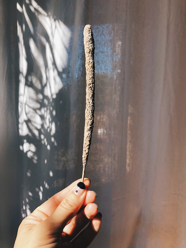 May include: A brown incense stick with a rough texture, held in a hand with black nail polish.