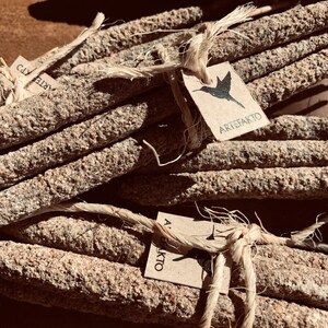 May include: A bundle of natural incense sticks tied together with twine. The sticks are made of a rough, textured material and have a brown and grey colour. The twine is a natural beige colour. A small paper tag with a hummingbird graphic and the text "ARTEFAKTO" is attached to the twine.