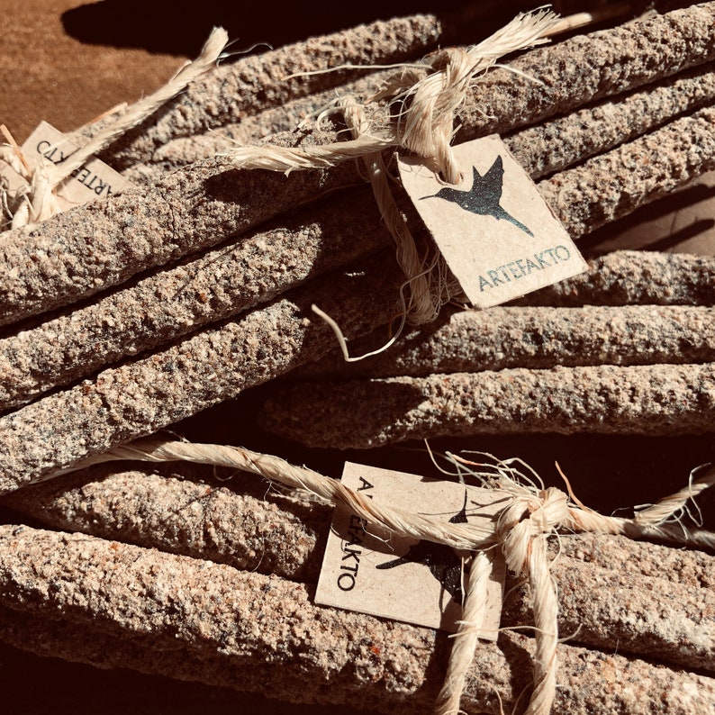 May include: A bundle of natural incense sticks tied together with twine. The sticks are made of a rough, textured material and have a brown and grey colour. The twine is a natural beige colour. A small paper tag with a hummingbird graphic and the text "ARTEFAKTO" is attached to the twine.