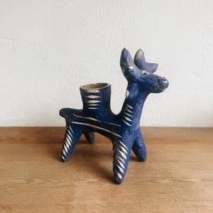 May include: A blue ceramic animal figurine with a hollow cylindrical opening on its back. The figure features white incised leaf-like patterns along its legs, body, and neck. It has four legs, a pointed tail, and a head with two small horns.