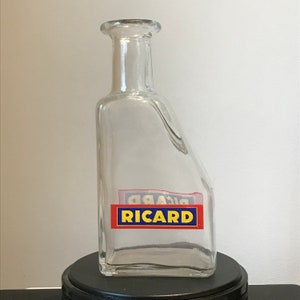 Puede incluir: Una botella de vidrio transparente con una forma única y una etiqueta roja y azul que dice "RICARD".