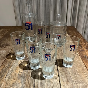 May include: A collection of clear glass bottles and glasses with the number "51" printed in blue and orange. The set includes two bottles and six glasses, all arranged on a wooden surface.