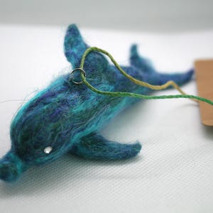 May include: A handmade felt dolphin in shades of teal and blue, with a small silver ring attached. The dolphin has a small, clear gem for an eye and a green and yellow string. A brown tag is attached.