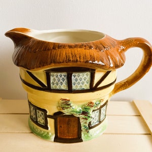 May include: A ceramic jug shaped like a cottage with a brown thatched roof, yellow walls, and brown trim. The jug has a brown handle and a white interior.