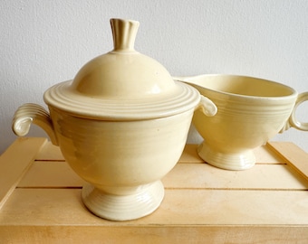 May include: A cream-colored ceramic sugar bowl with a lid and a matching creamer. The sugar bowl has a pedestal base and curved handles. The creamer has a handle and a spout. Both pieces are on a light wood surface.