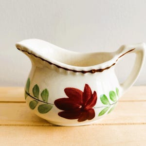 May include: A white ceramic creamer with a brown rim and a floral design. The design features a large red flower with green leaves.