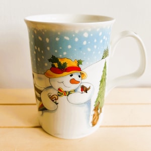 May include: A white ceramic mug with a snowman design. The snowman is wearing a yellow hat with red trim and a green holly sprig. A small bird is perched on the snowman's arm. The background is a blue and white winter scene.