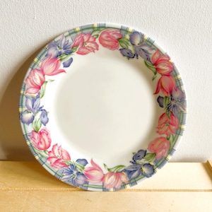 Peut inclure: Une assiette ronde en céramique blanche avec un motif floral. La bordure présente des tulipes roses et des iris violets, sur un motif à carreaux vert et bleu. L'assiette est adaptée pour servir des aliments.