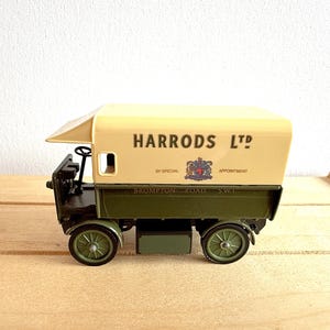 May include: A vintage toy truck with a cream-colored top and olive green bed. The truck features the text "HARRODS LTD" and a crest. The wheels are olive green, and the truck is set on a wooden surface.