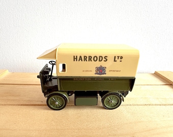 Puede incluir: Un camión de juguete vintage con techo crema y caja verde oliva. El camión presenta el texto "HARRODS LTD" y un escudo. Las ruedas son verde oliva, sobre una superficie de madera.