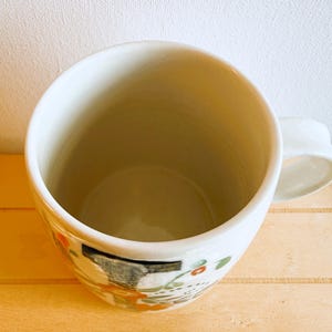Op de afbeelding: Een witte keramische mok met een afgeronde body en een handvat. De mok heeft een geschilderd ontwerp met oranje, groene en zwarte accenten. De mok staat op een lichtgekleurd houten oppervlak.