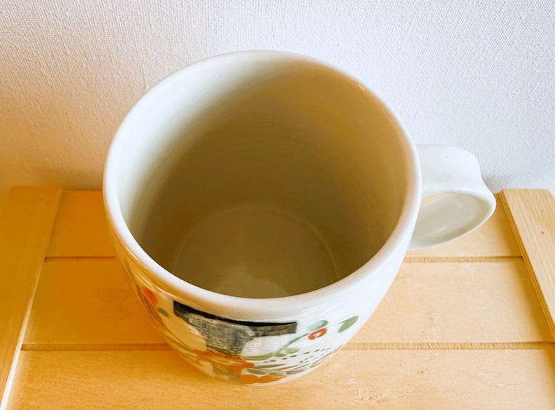 Op de afbeelding: Een witte keramische mok met een afgeronde body en een handvat. De mok heeft een geschilderd ontwerp met oranje, groene en zwarte accenten. De mok staat op een lichtgekleurd houten oppervlak.