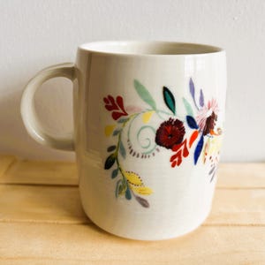 Op de afbeelding: Witte keramische mok met een gebogen handvat, versierd met een kleurrijk bloemenontwerp. De bloemen hebben rode, gele, blauwe en groene tinten. De mok staat tegen een lichte achtergrond.