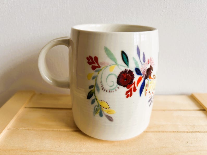Op de afbeelding: Witte keramische mok met een gebogen handvat, versierd met een kleurrijk bloemenontwerp. De bloemen hebben rode, gele, blauwe en groene tinten. De mok staat tegen een lichte achtergrond.