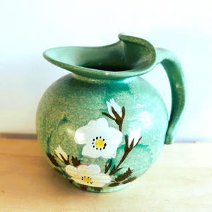 May include: A green ceramic pitcher with a floral design. The pitcher has a wide mouth and a handle. The flowers are white with yellow centers and brown stems.