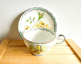 May include: A white teacup and saucer set with a floral design. The cup and saucer have a light blue scalloped edge and gold trim. The floral pattern features yellow and green flowers.