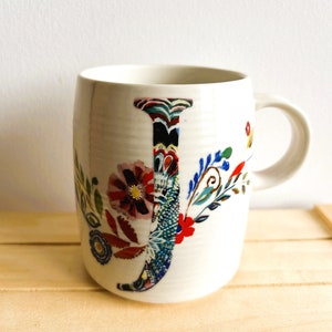 Op de afbeelding: Witte keramische mok met een groot, kleurrijk bloemen "J" ontwerp. De letter is gevuld met ingewikkelde patronen en omgeven door extra bloemen- en bladmotieven in rood, blauw en groen. De mok heeft een gebogen vorm en een wit handvat.