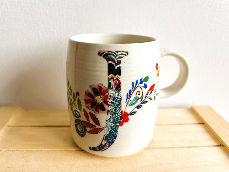 Op de afbeelding: Witte keramische mok met een groot, kleurrijk bloemen "J" ontwerp. De letter is gevuld met ingewikkelde patronen en omgeven door extra bloemen- en bladmotieven in rood, blauw en groen. De mok heeft een gebogen vorm en een wit handvat.