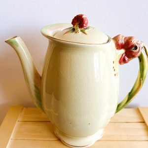 May include: A cream-coloured ceramic teapot with a floral design. The teapot has a lid with a pink and green floral design and a handle shaped like a flower with red petals.