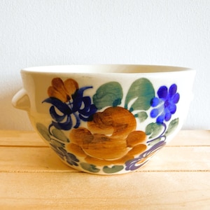 Könnte beinhalten: Eine weiße Keramikschale mit floralem Design. Die Schale hat braune, blaue und grüne Blumen. Die Schale hat zwei kleine Griffe und steht auf einer Holzoberfläche.