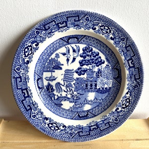 Puede incluir: Un plato de porcelana azul y blanco con un diseño tradicional de sauce. El plato presenta un paisaje estilizado con un puente, una pagoda y un sauce.