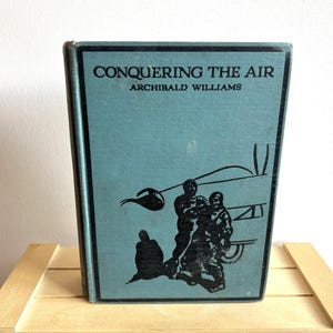May include: A blue hardcover book titled "CONQUERING THE AIR" by ARCHIBALD WILLIAMS. The cover features a black and white illustration of three people in a biplane.