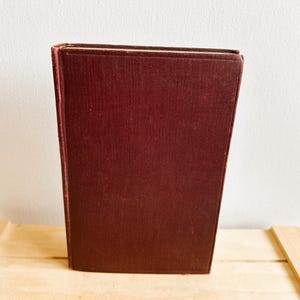 May include: A maroon-colored book with a textured cover stands upright on a wooden surface. The book's spine is visible, and the pages are slightly yellowed. The background is a plain white wall.