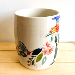Op de afbeelding: Een witte keramische beker met een bloemenpatroon. De cilindrische beker is versierd met kleurrijke bloemen, bladeren en andere botanische elementen. Het ontwerp bevat rode, oranje, blauwe en groene accenten.