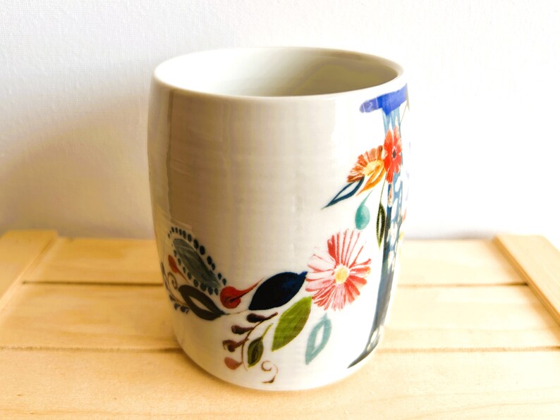 Op de afbeelding: Een witte keramische beker met een bloemenpatroon. De cilindrische beker is versierd met kleurrijke bloemen, bladeren en andere botanische elementen. Het ontwerp bevat rode, oranje, blauwe en groene accenten.