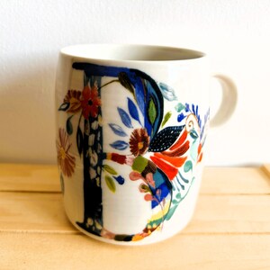 Op de afbeelding: Witte keramische mok met een groot, kleurrijk bloemmotief. Het ontwerp bevat de letter "F" in donkerblauw, met rode, oranje en blauwe bloemen en bladeren. De mok heeft een gebogen vorm en een oor.
