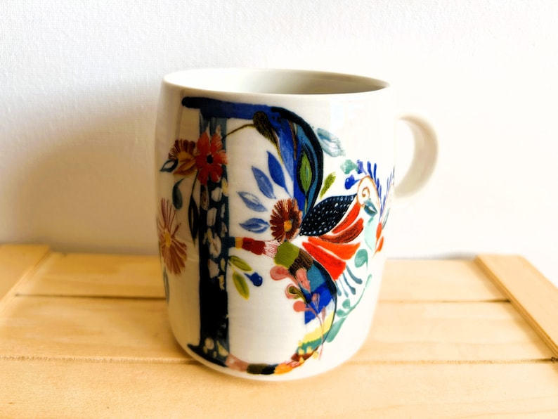Op de afbeelding: Witte keramische mok met een groot, kleurrijk bloemmotief. Het ontwerp bevat de letter "F" in donkerblauw, met rode, oranje en blauwe bloemen en bladeren. De mok heeft een gebogen vorm en een oor.