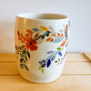 Op de afbeelding: Witte keramische beker met een bloemenontwerp. De beker heeft een kleurrijk patroon van bloemen en bladeren in tinten rood, oranje, blauw en groen. Het ontwerp wikkelt zich om de cilindrische vorm van de beker.