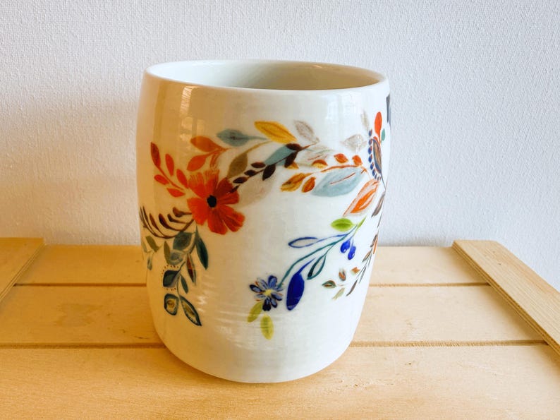 Op de afbeelding: Witte keramische beker met een bloemenontwerp. De beker heeft een kleurrijk patroon van bloemen en bladeren in tinten rood, oranje, blauw en groen. Het ontwerp wikkelt zich om de cilindrische vorm van de beker.