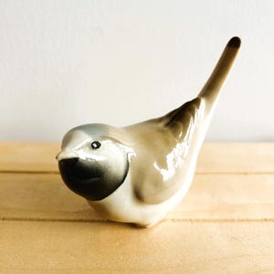 Könnte beinhalten: Eine Keramik-Vogelfigur mit brauner und grauer Glasur. Der Vogel hat einen langen Schwanz und einen kleinen Schnabel. Die Figur steht auf einer Holzoberfläche.