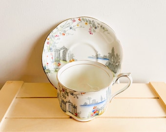 May include: A white teacup and saucer with a floral pattern and a gold rim. The pattern features a gate, flowers, and a landscape scene.