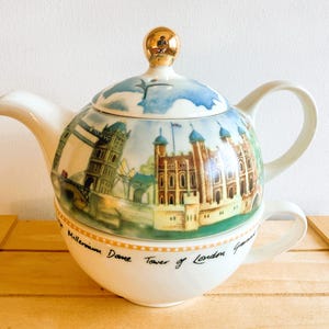 Puede incluir: Un juego de té de cerámica blanca que consiste en una tetera apilada sobre una taza. La tapa de la tetera tiene un pomo dorado. El exterior muestra una ilustración colorida de monumentos de Londres con texto impreso en la base.