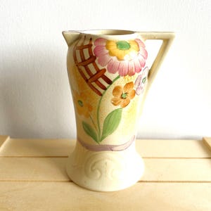 May include: A cream-colored ceramic pitcher with a floral design. The pitcher has a wide mouth and a handle. The flowers are painted in shades of pink, yellow, and orange.