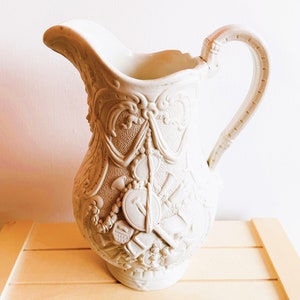 Puede incluir: Una jarra de cerámica blanca con un diseño ornamentado que presenta un patrón floral y un diseño en relieve de un escudo y otros objetos. La jarra tiene un asa y una boca ancha.