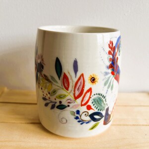Op de afbeelding: Een witte keramische beker met een kleurrijk bloemmotief. Het ontwerp bevat rode, blauwe, groene en gele bloemen en bladeren. De beker is cilindrisch en staat op een lichtbruin houten oppervlak.