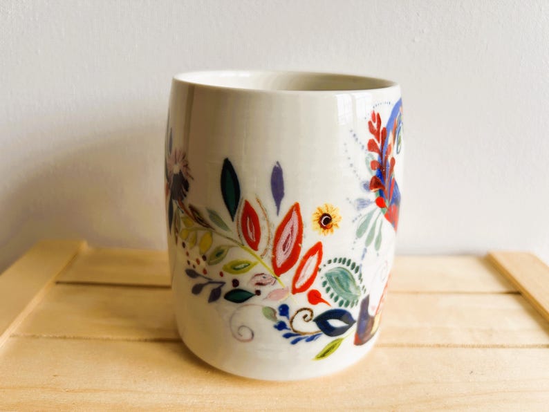 Op de afbeelding: Een witte keramische beker met een kleurrijk bloemmotief. Het ontwerp bevat rode, blauwe, groene en gele bloemen en bladeren. De beker is cilindrisch en staat op een lichtbruin houten oppervlak.