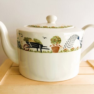 Charming vintage Holly Cottage oval porcelain teapot. Creamy white glaze with wonderful border in a garden-theme. Pattern includes plants, wheelbarrows, watering cans, flowers, birds and even a lawn roller.