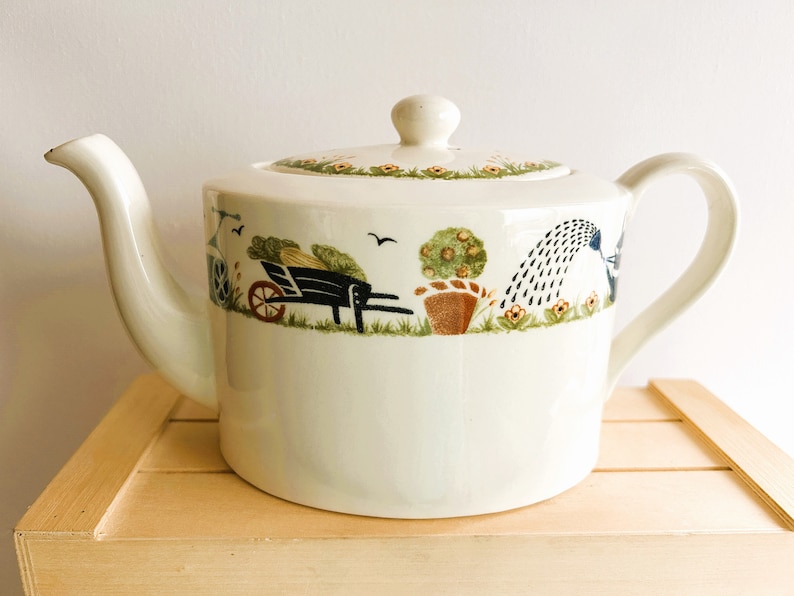 Charming vintage Holly Cottage oval porcelain teapot. Creamy white glaze with wonderful border in a garden-theme. Pattern includes plants, wheelbarrows, watering cans, flowers, birds and even a lawn roller.
