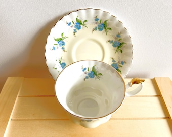 May include: A vintage teacup and saucer set. The cup and saucer are cream-colored with a delicate floral pattern of blue flowers and green leaves. The rims are trimmed with gold. The cup has a gold handle.
