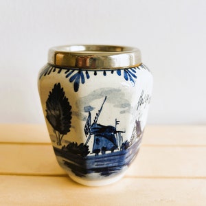 以下が含まれることがあります： 銀色の縁が付いた、青と白のセラミックジャー。オランダの風車デザインが特徴です。ジャーは円筒形で、わずかにフレアしたベースがあり、風景が描かれています。