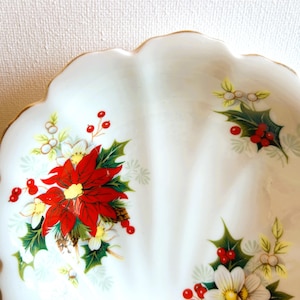 K&ouml;nnte beinhalten: Eine wei&szlig;e Keramik-Servierplatte mit gewelltem Rand und goldener Einfassung. Die Platte ist mit einem Blumenmuster verziert, das rote Weihnachtssterne, gr&uuml;ne Stechpalmenbl&auml;tter und wei&szlig;e Beeren zeigt.