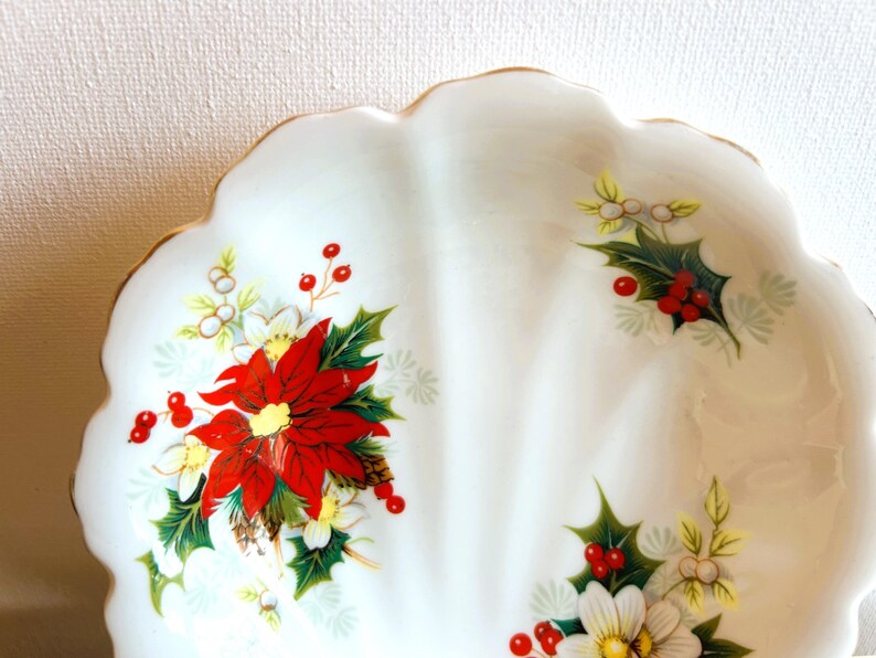 K&ouml;nnte beinhalten: Eine wei&szlig;e Keramik-Servierplatte mit gewelltem Rand und goldener Einfassung. Die Platte ist mit einem Blumenmuster verziert, das rote Weihnachtssterne, gr&uuml;ne Stechpalmenbl&auml;tter und wei&szlig;e Beeren zeigt.