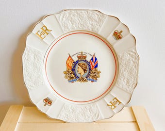 May include: White decorative plate with a scalloped edge, featuring a central emblem with a profile of a woman, flags, and the text "Coronation June 1953". Gold "ER" initials and crown details adorn the plate.